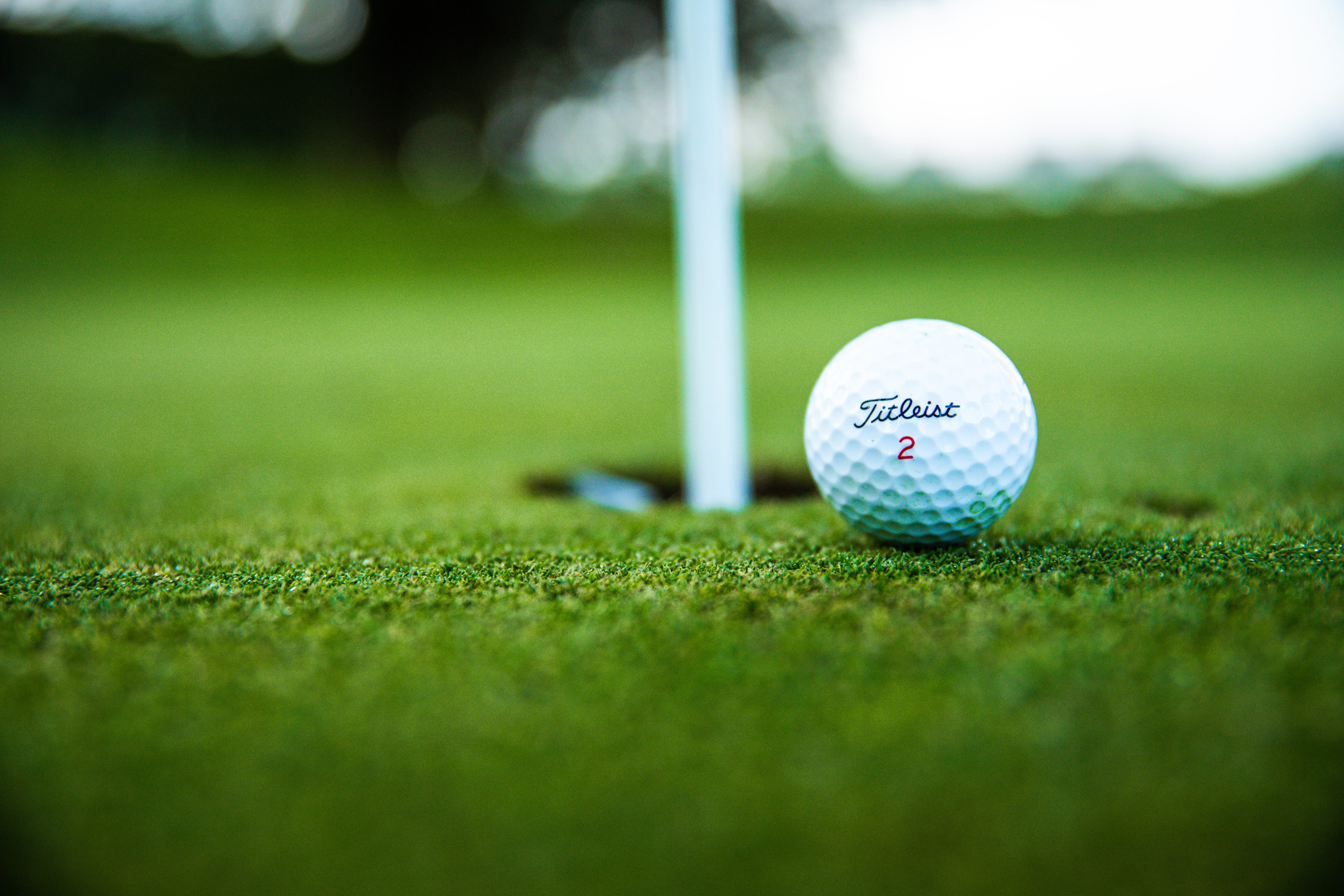 A white golf ball sits on the green while it balances on the edge of a whole in the ground with a white pole sticking up