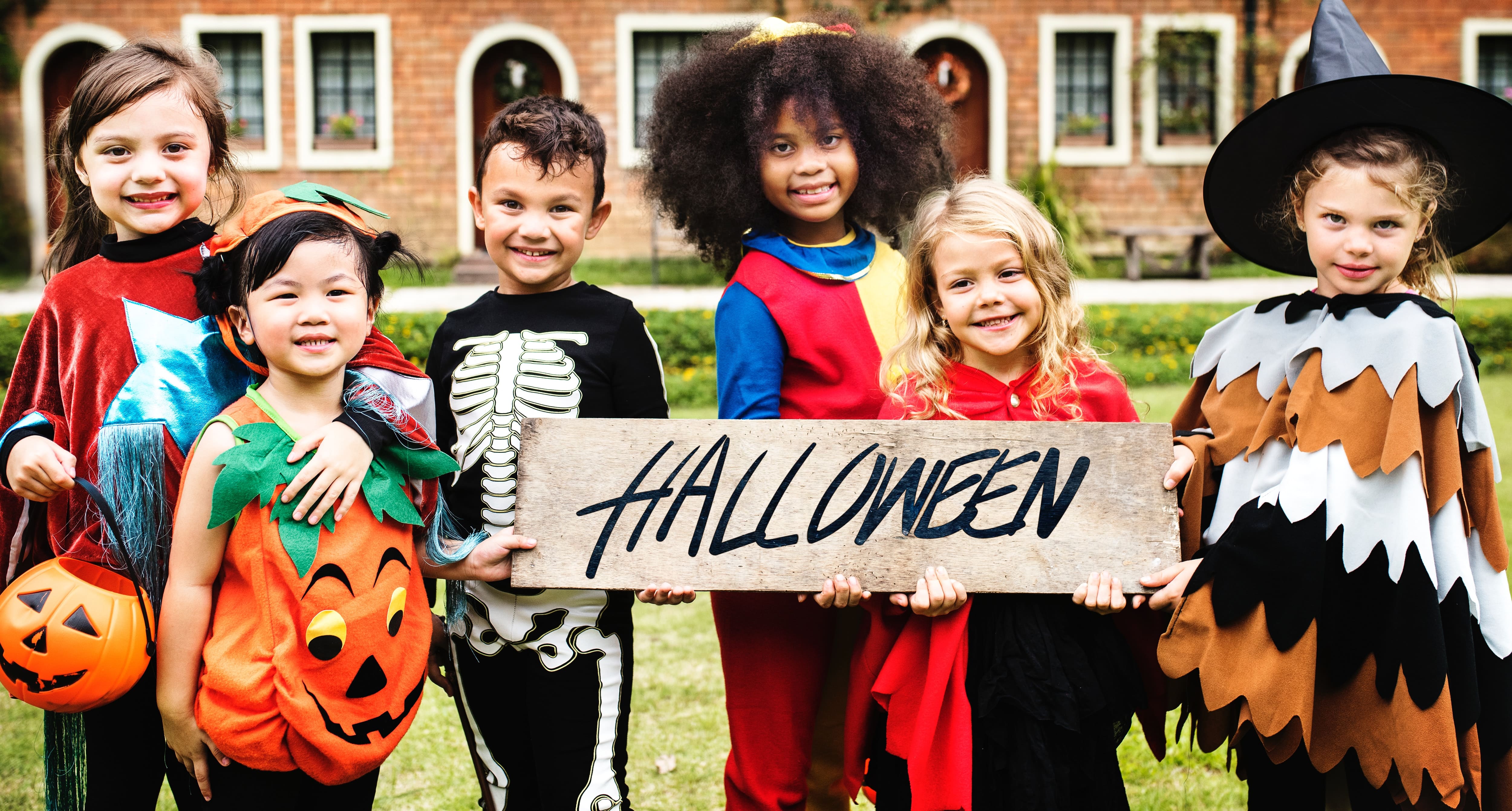 A group of six children dressed in various Halloween costumes stand outside holding a wooden sign that says "Halloween"